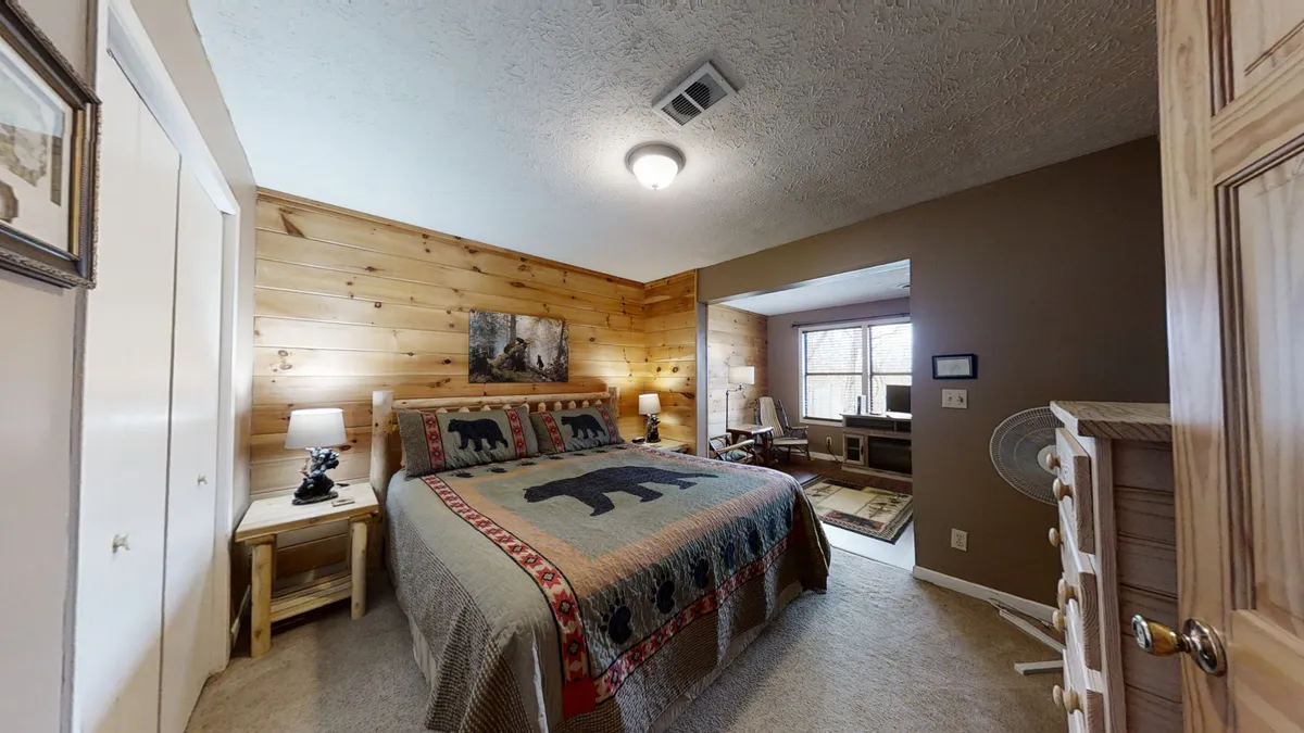A warmly lit bedroom with a queen-sized bed dressed in a charming bear motif quilt, wood-paneled walls, framed artwork, and matching nightstands with lamps. The setting is inviting and cozy, perfect for a restful night's sleep.