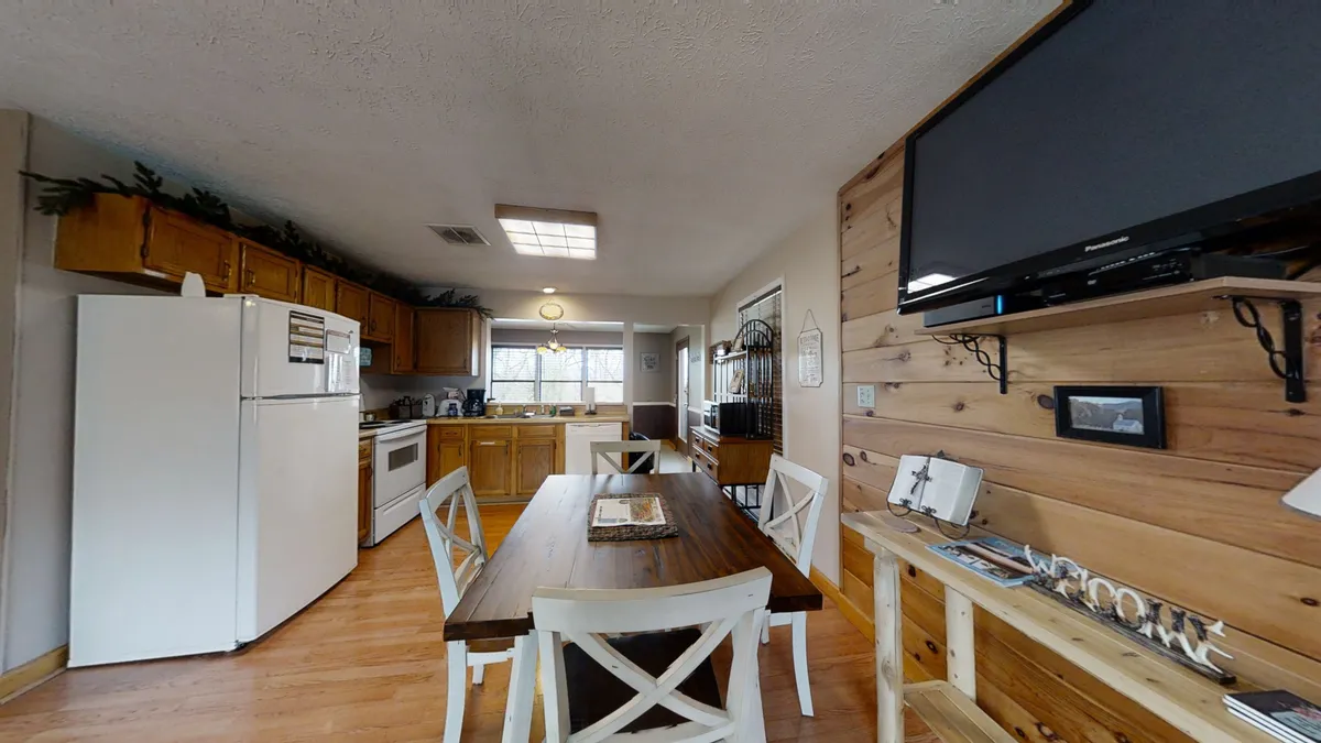 This image showcases a cozy and inviting dining area with a wooden dining table set for four, kitchen appliances visible in the background, and charming wood wall decorations. The layout highlights a seamless transition from cooking to dining, making it clear this is a space for both meal preparation and gathering.