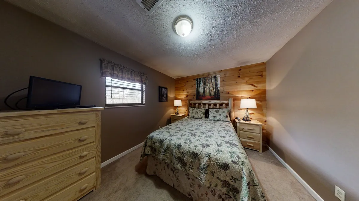 A cozy bedroom featuring a king-sized bed with patterned bedding, wooden accent wall, matching nightstands with soft lamps, and a flat-screen television facing the bed. The space feels warm, inviting, and perfect for relaxing after a long day.