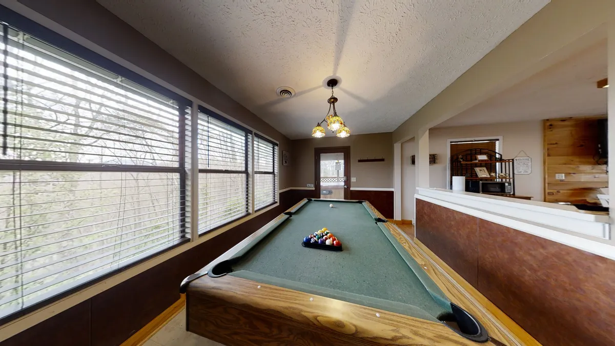 A warmly lit recreation room features a pool table set near expansive windows, allowing natural light to pour in. A modern chandelier hangs overhead, and the room is designed for gathering, with ample space for families to mingle, play games, and enjoy each other's company.