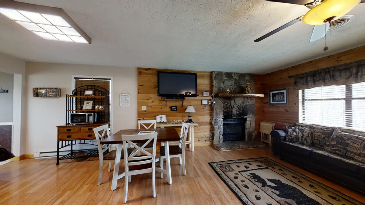 A warm and homely scene featuring a wooden dining table with four chairs, a stone fireplace, a cozy sofa with patterned upholstery, and a television. Decorative items on the mantel add character. The setting evokes the experience of gathering for meals, sharing stories, and relaxing together after a day of adventures.