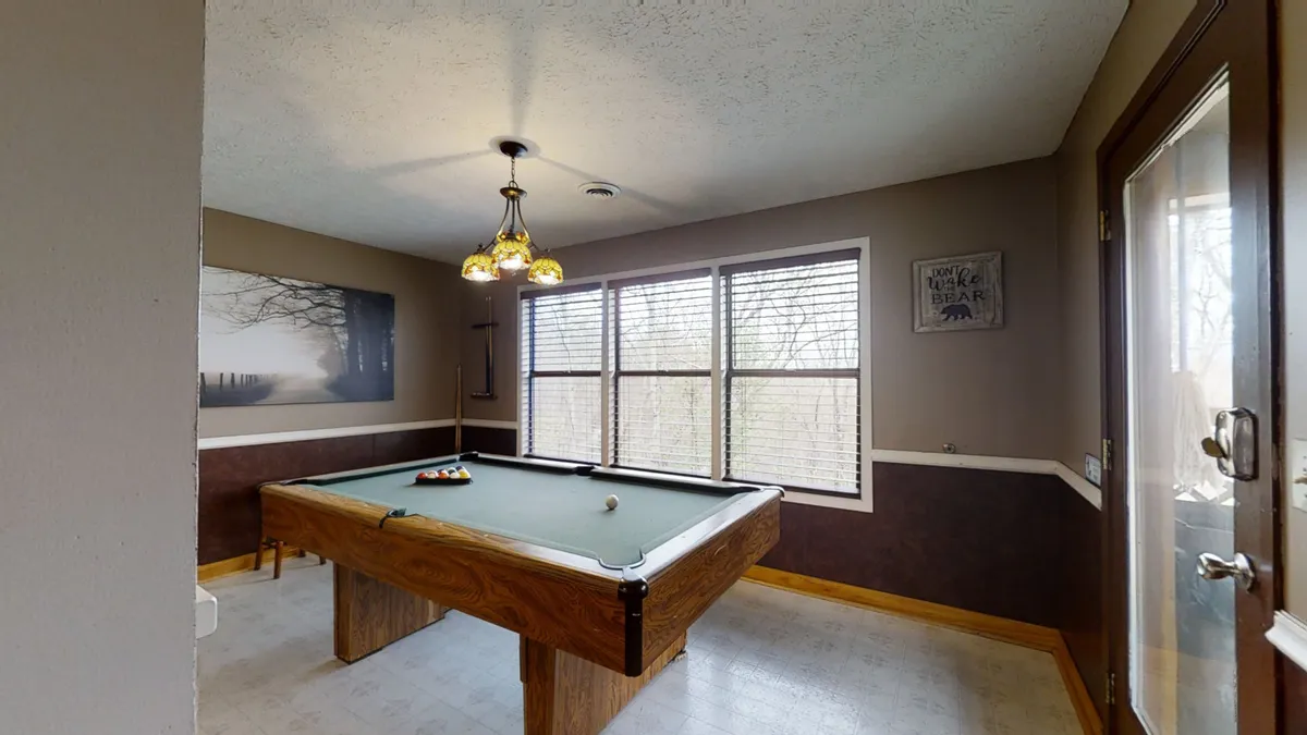 This image captures a lively in-cabin scene centered on a pool table, surrounded by large windows that let in natural light. Friends are playing billiards and socializing under a prominent hanging light fixture, evoking a warm, communal atmosphere ideal for game nights.
